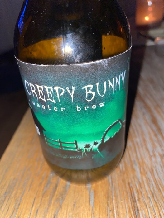 Flaska med etikett "CREEPY BUNNY easter brew", mörkt öl, staket, mörk bakgrund, träyta.
