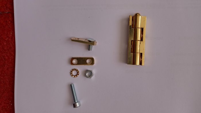 Beslag och komponenter för montering: skruvar, brickor, metallplatta och gångjärn på vit bakgrund.