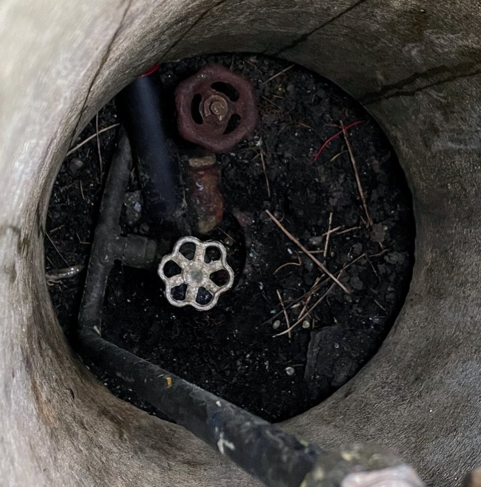 En öppen brunnslock, vattenrör, ventiler, smuts, rörkopplingar inuti en betongbrunn.