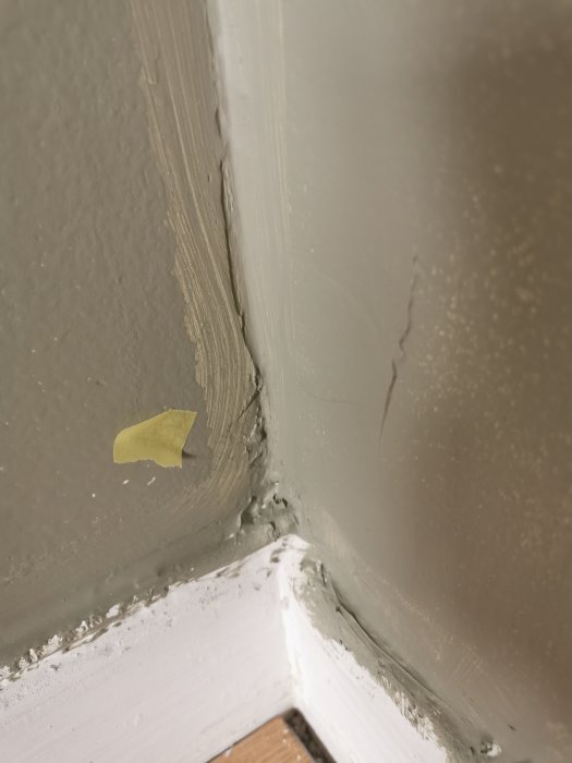 Vägghörn med spackling, sprickor, en bit tejp och vit färg på lister. Behov av underhåll synligt.