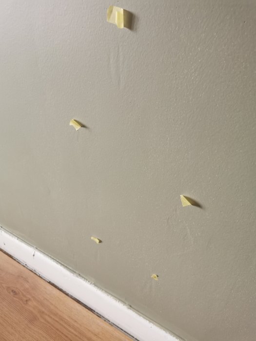 En vägg med maskeringstejpbitar ovanför en vit golvlist och ljust trägolv.