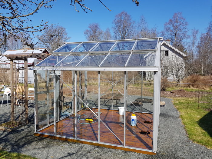 Ett växthus med trägolv och aluminiumramar på en solig dag med blå himmel; träd och ett hus i bakgrunden.