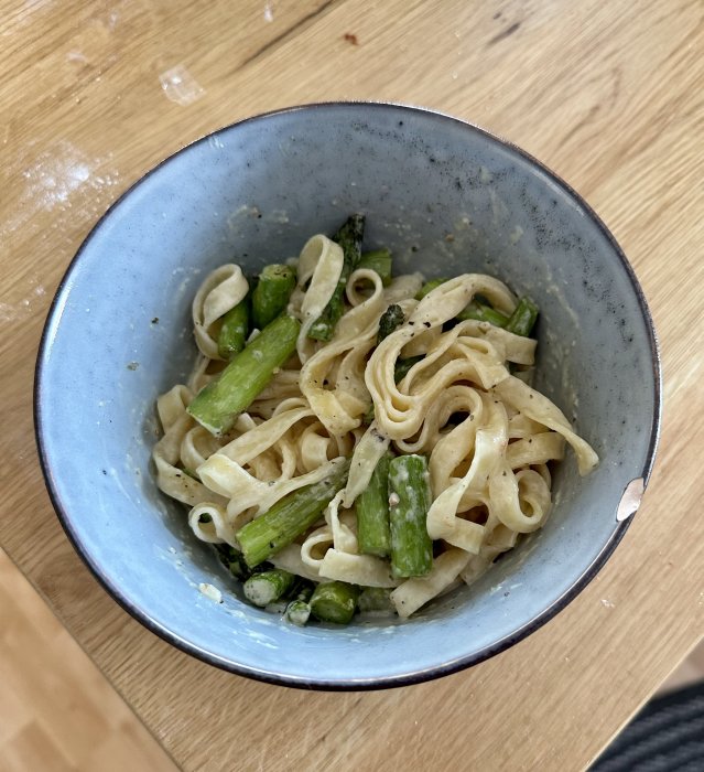 En skål med pasta och gröna sparrisbitar på ett träbord.