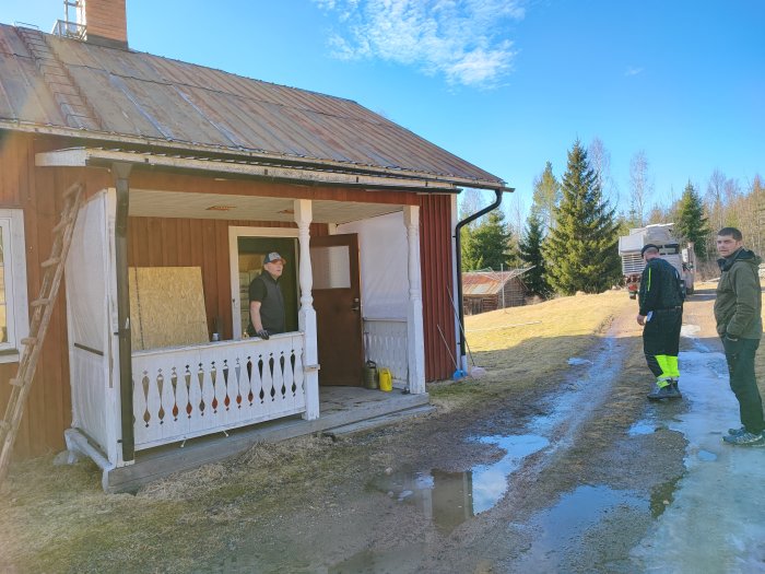 Två män utanför ett äldre hus med snösmältning och lerig mark, stegar, delvis täckt fönster, soligt väder.