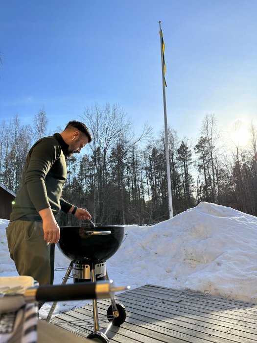 Man grillar utomhus på trädäck, omgiven av snö, under en klar himmel, nära svensk flagga.