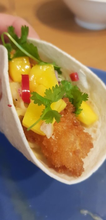 Taco med panerad fisk, mango, rödlök, koriander, hålls i hand. Närbild, matpresentation, otydlig bakgrund.