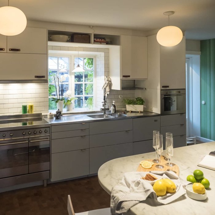 Modernt kök med grå skåp, vita kakelväggar, marmorö och rostfria vitvaror. Dekorerat bord med citroner, glas.