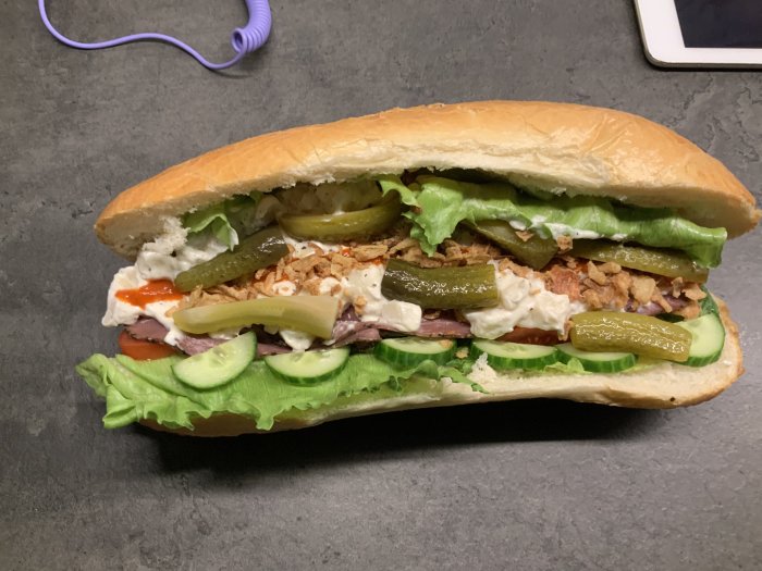 Ett överfyllt baguette-sandwich med sallad, tomat, gurka, pickles, vit sås och röd sås. På ett bord.