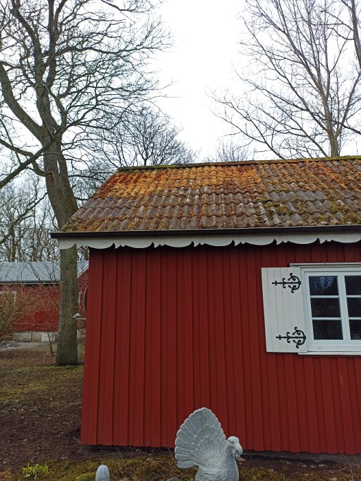 Rött trähus, tegeltak med mossa, fönsterluckor, träd, dekorativ trädgårdsturky i förgrunden.