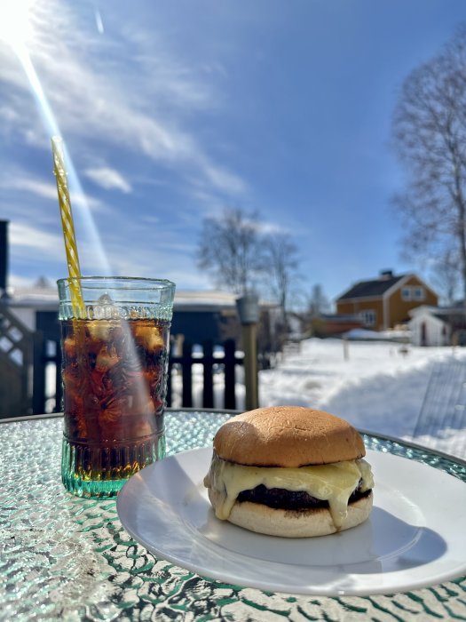 Ostburgare på tallrik, iskall dryck med sugrör, soligt väder, snö, utemöbler, klarblå himmel.