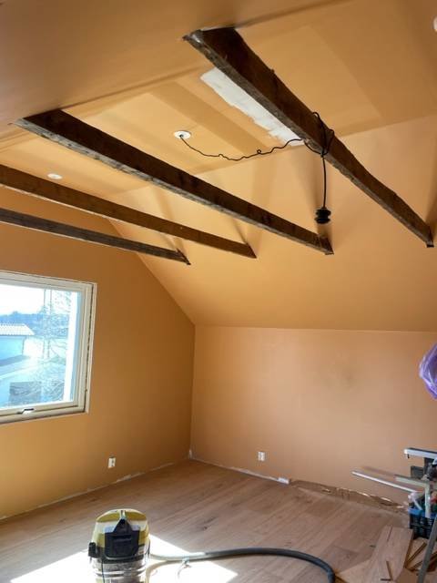 Rum under renovering, träbjälkar i taket, hängande lampa, fönster, arbetsmaterial på golvet.