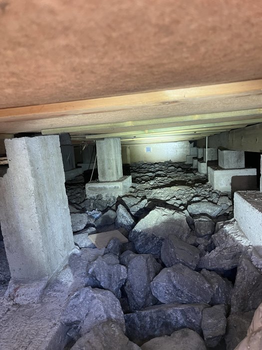 Krypgrund under hus med stenar, betongpelare och träbjälkar. Diffust ljus, installationer synliga i bakgrunden.