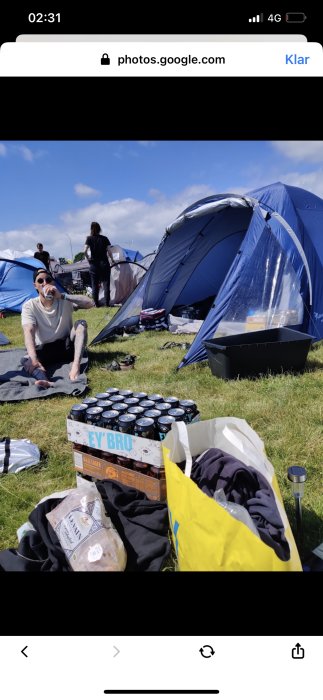 Person sitter bland tält på festival, dricker öl, avslappnad stämning, soligt väder, campingutrustning och förnödenheter runtomkring.