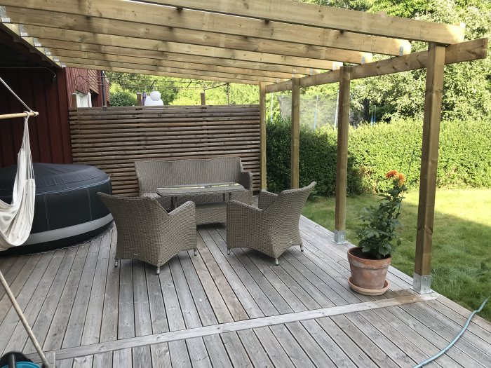 Trädäck med rottingmöbler, pergola, hängmatta, blomkruka och grönt gräs, nära ett rött hus.