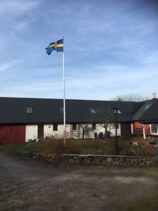 Svensk flagga vajar framför lantliga byggnader under en klarblå himmel.