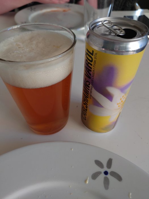 Ett glas öl, en öppnad burk, tom tallrik med smulor, på ett bord.