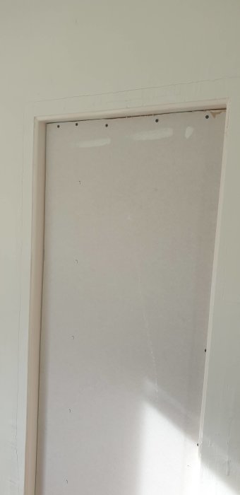En ofullständig dörröppning med täckplatta och synliga skruvhål mot en vit vägg.