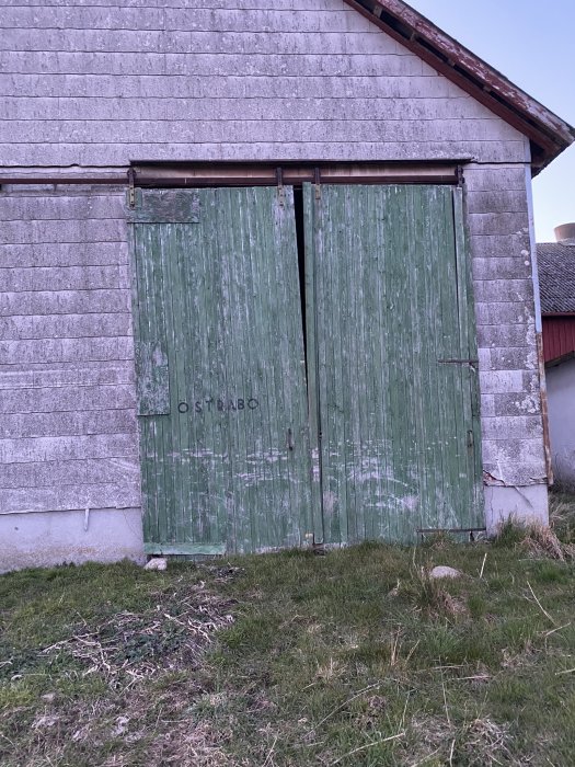 Sliten grön ladugårdsdörr, asbestcementskivor, "Ostabö" målat överst, skymning, gräs framför, förfall.