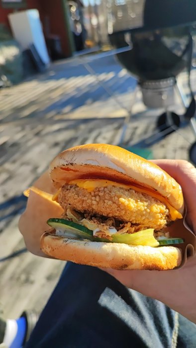 En hand håller en kycklingburgare med ost och grönsaker, suddig bakgrund av stadsmiljö.