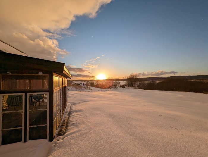 Solnedgång, snötäckt landskap, fotspår i snö, gammal byggnad, klart väder, moln på himlen.