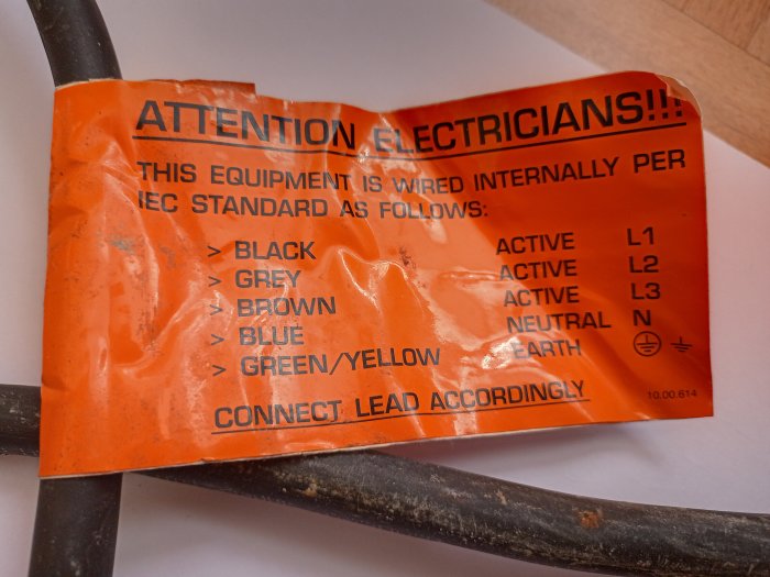 Orange varningsetikett för elektriker med ledningsinstruktioner, sliten och ligger på kabel.