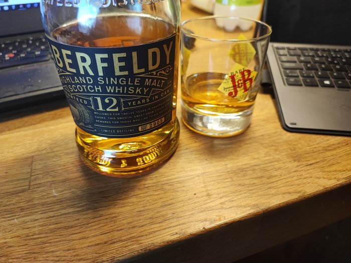 Aberfeldy whiskyflaska, glas med whisky, laptop på trädskrivbord. Arbete och avkoppling kombineras.
