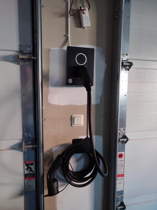 Elbilsladdningsstation installerad på en vägg bredvid en garageport, med upphängd laddningskabel.