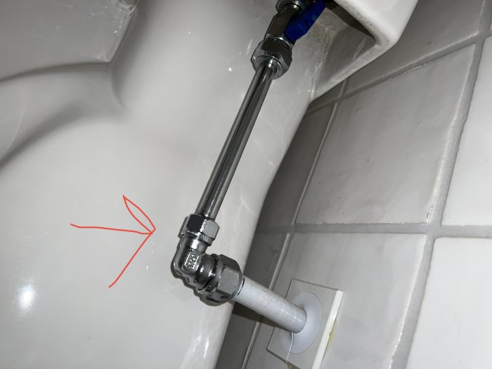 Toalett med ansluten vattenledning, blå märkning, vit kakelvägg, röd pil pekar mot hörn.