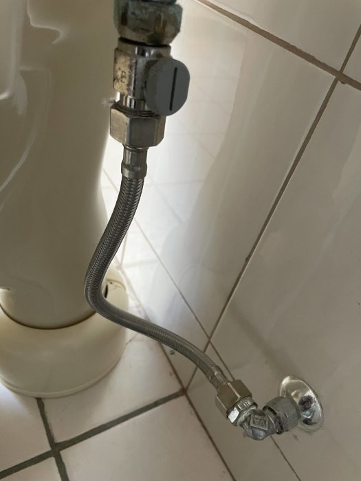 Metallisk flexibel slang ansluter en vit toalett till en vattenventil mot kaklad vägg.