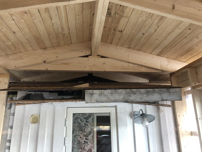 Träklädd veranda, vit dörr, lampa, skador i taket som visar isolering, renoveringsbehov.