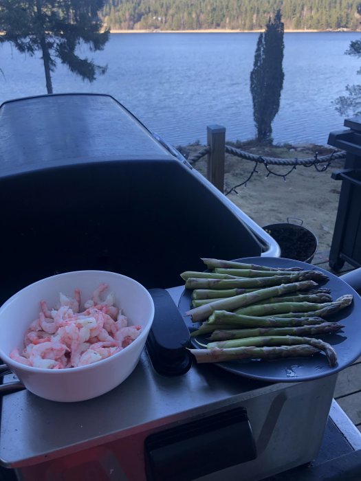 Matlagning utomhus: skaldjur och sparris på grillen vid en sjö med träd.