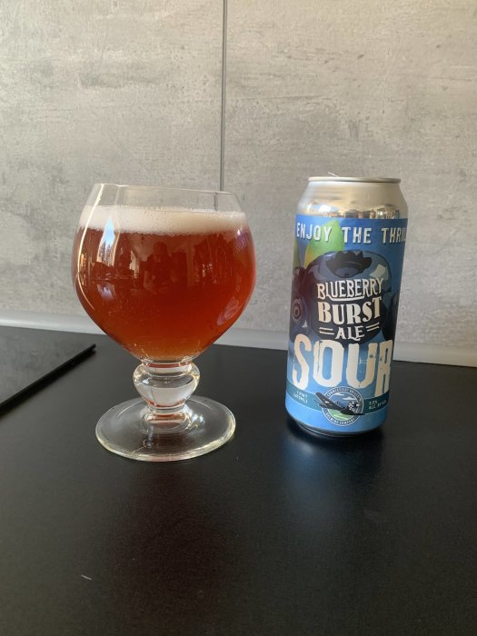 Ett glas med orange dryck bredvid en burk märkt "Blueberry Burst Sour Ale".