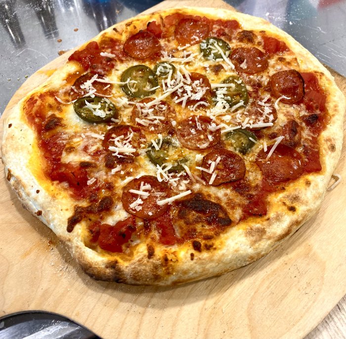 Nybakad pizza med pepperoni, jalapeños och riven ost på träskärbräda.