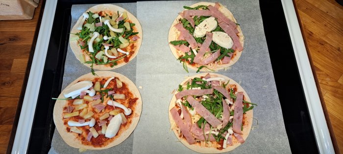 Olagade pizzor på bakplåtspapper med skinka, ananas, lök, mozzarella, rucola och vitlök redo för ugnen.