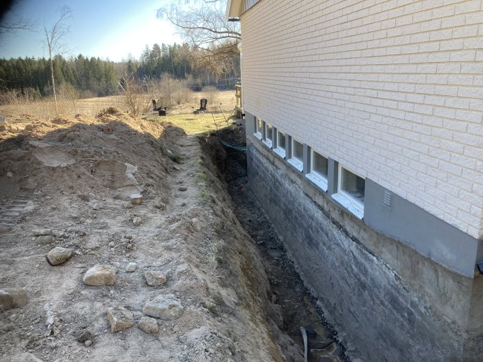 Grävd schakt vid husgrund, brun jord, stenar, källarfönster, skyddsnät, träd i bakgrunden.