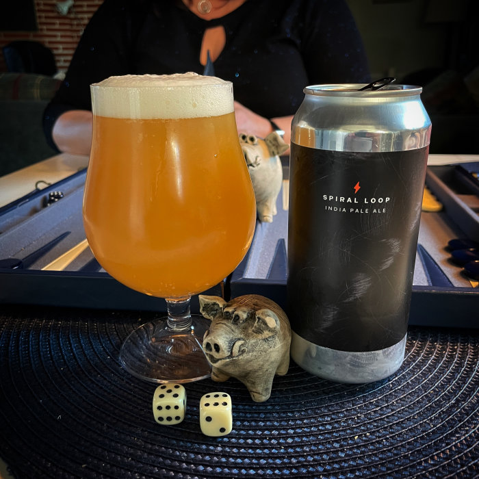 Öl i glas, burk "Spiral Loop IPA", tärningar, svinfigurin, backgammonbräde i bakgrunden, del av en person.