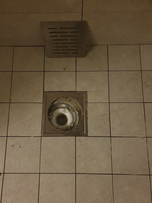 Golv med plattor, brunnen öppen med synlig avlopp, ventilation vid sidan. Behöver underhåll eller rengöring.