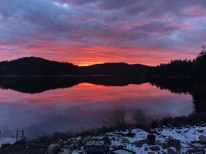 Solnedgång med levande färger reflekterar över stilla vatten vid skogsbeklädda bergen och snöfläckad mark.