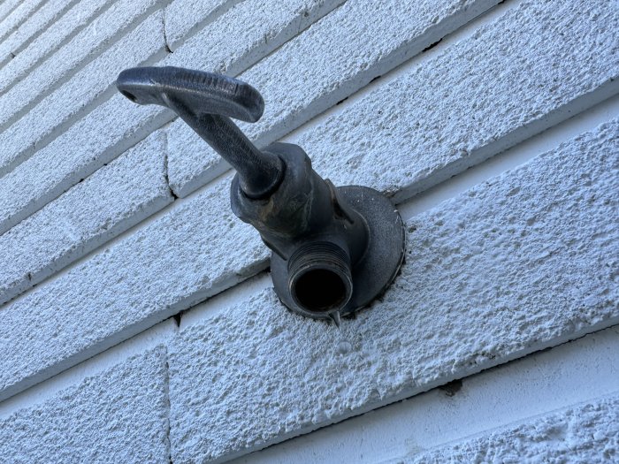 Väggmonterad, gammal, rostig vattenkran på en vit träsidovägg av ett hus.