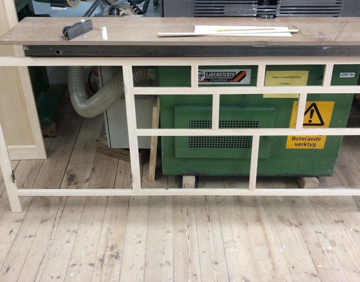 En arbetsbänk i en verkstad med en grön maskin och varningsskyltar om roterande verktyg.