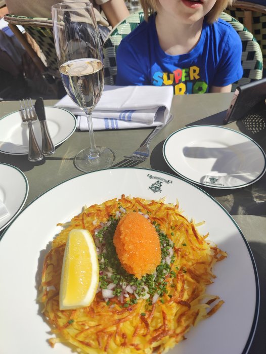 En maträtt med potatis, löjrom, rödlök, persilja och citron serveras utomhus med ett glas skumpa. En person syns.