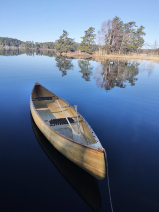 Kanot på spegelblank sjö, träd speglar sig i vattnet, klarblå himmel, lugn och fridfull.