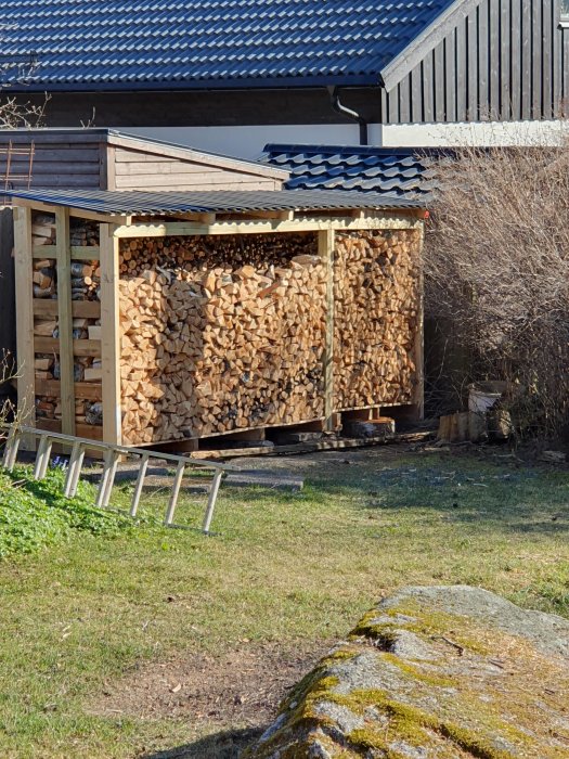 Vedskjul fullt med vedstack, grönt gräs, stege, träbyggnad i bakgrunden, dagtid, soligt, utomhus.