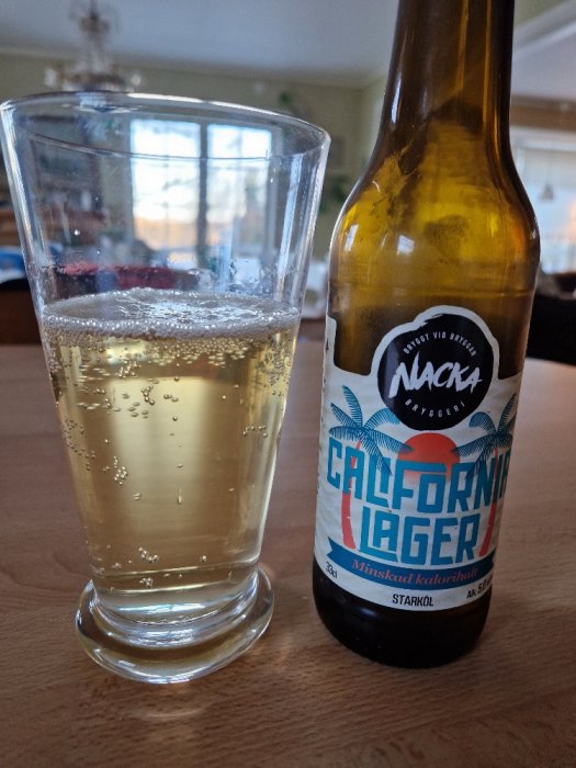 Ett glas med ljus dryck bredvid en ölflaska märkt "California Lager" från Nacka Bryggeri på ett bord.