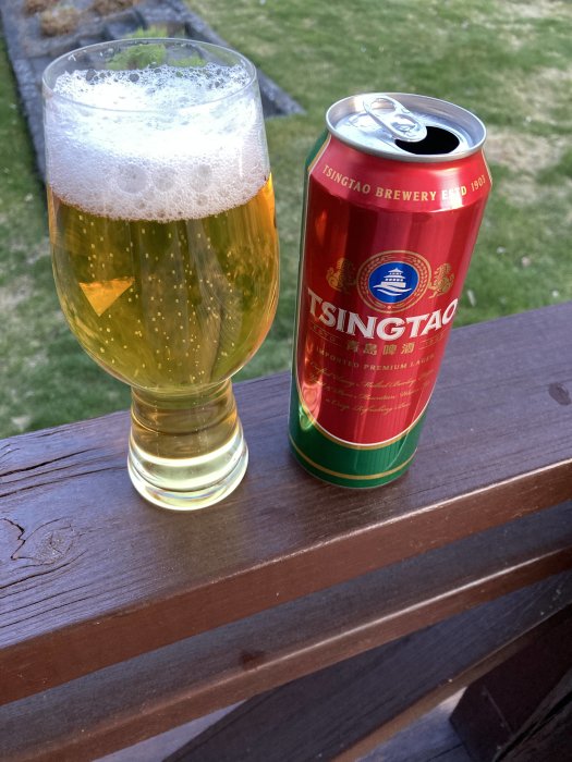 Tsingtao ölburk bredvid glas fyllt med öl, utomhus på träledning, gräsmatta i bakgrunden.