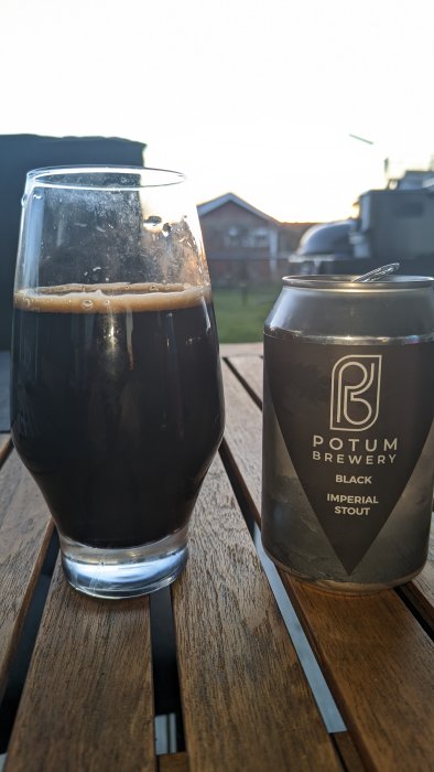 En mörk öl i glas bredvid dess burk, Potum Brewery Imperial Stout, utomhus på träbord.