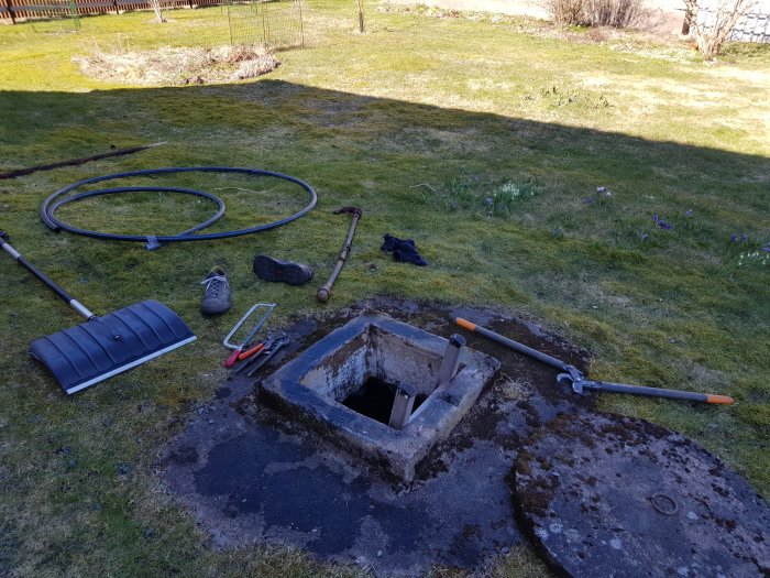 En öppen brunn med lock bredvid, verktyg och utrustning på gräset. Vårblommor syns i trädgården.