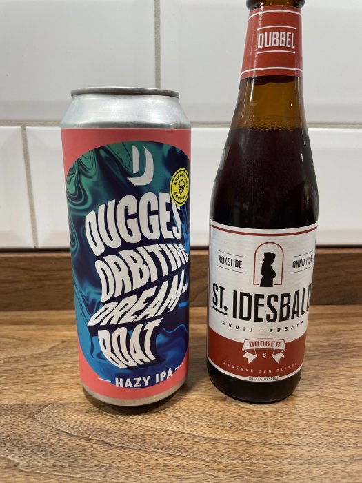 Två ölflaskor, en burk Hazy IPA och en Dubbel, på en träyta mot kaklad vägg.