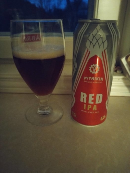 Ett glas med mörkröd öl bredvid en burk med texten "RED IPA", fönster och skymning i bakgrunden.