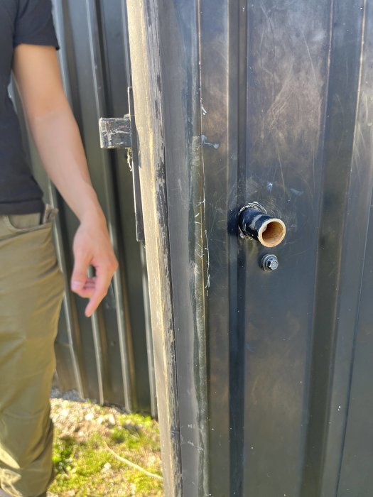 Närbild på svart dörr, trasigt lås, person pekar på mekanism, dagtid, utomhus, synlig gångjärn, blek arm.